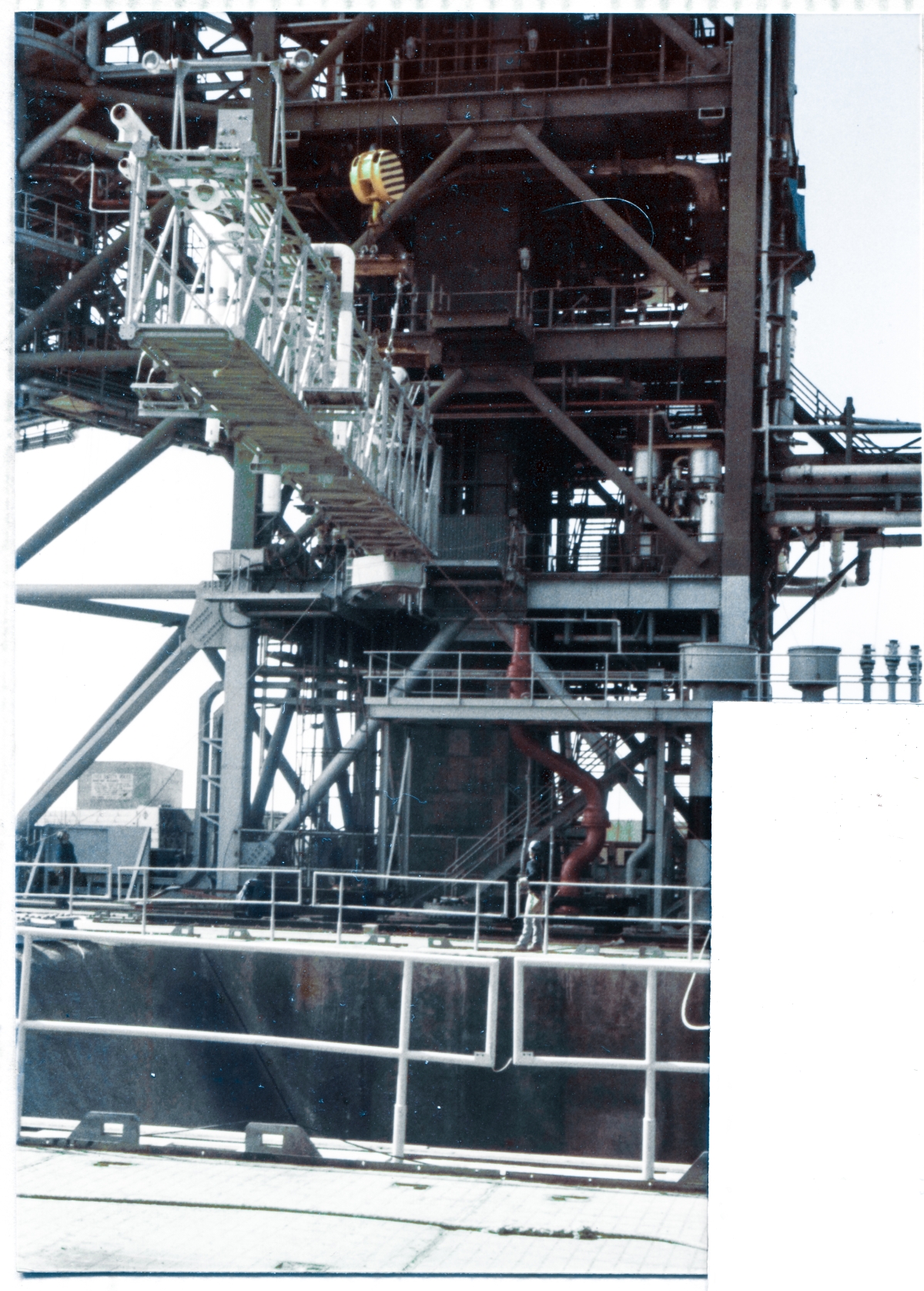 Image 103. At Space Shuttle Launch Complex 39-B, Kennedy Space Center, Florida, the GOX Arm is being lifted by the Hammerhead Crane on top of the Fixed Service Structure, and has reached an intermediate position during the Lift, where Union Ironworkers working for Ivey Steel Erectors are using tag lines attached to either end of the Arm to rotate it around into an orientation more closely-aligned with the final orientation it will assume when it gets bolted to the top of the FSS. Note: The blocked-out area in the photograph represents an overlapping print photograph on the same page in the original photo albums which these images were stored in, which was not removed from its overlapping position in order to keep these decades-old artifacts as protected as possible from any damage which might have occurred had they been removed or otherwise handled. Whenever it comes time to show the overlapping image, that will be done, and the entire photograph will be visible, but the portions of this and other photographs, which lay beneath, never got scanned, and therefore will never become visible in these photo essays. Overlap occurred due to insufficient room on the album pages, and in order to reduce visual confusion, the overlap was simply blocked out as you are seeing it here. Photo by James MacLaren.